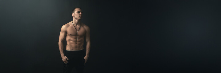 Fototapeta premium Muscular Man with Rosary on Dark Wide Banner with Copy Space