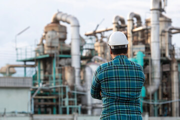 Industry Professional at the Factory: An engineer, dressed in a plaid shirt and safety hat, observes the complex machinery of a modern factory, embodying competence and expertise.