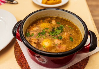 Close-up of żurek, a traditional Polish sour rye soup with sausage, potatoes, and fresh parsley, served in a rustic enamel pot on a wooden table in a restaurant setting.