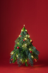 Small Christmas tree made of spruce branches decorated with red balls and glowing lights on red background creates festive holiday mood