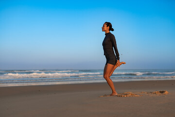 Sporty woman stretching quadriceps on sandy beach at sunrise, standing in active fitness pose near ocean, wearing black training outfit, focusing on flexibility, balance, and healthy morning routine.