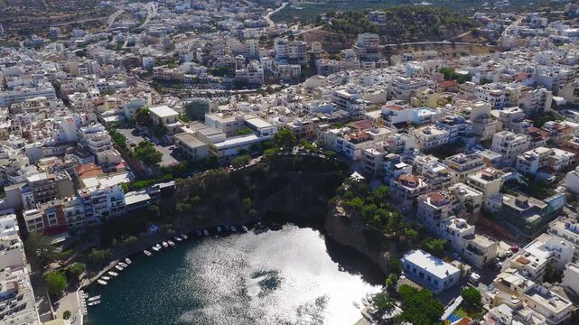 A slow sweeping aerial pan of Agios Nikolaos, Crete, showing white and pastel buildings, lush greenery, and rugged mountains under a clear sky.
