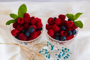 Two glass dessert bowls filled with ripe raspberries and blueberries, topped with fresh mint leaves on a white linen background. Perfect for healthy eating, summer fruit and dessert themes