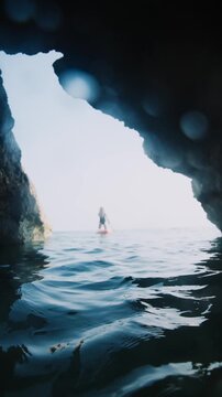 A woman is paddle boarding in the ocean. The water is calm and the sky is blue. The woman is in the middle of the water and the rock formation is in the background