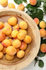 Fresh ripe apricots in bowl with water and green leaves on white table, flat lay