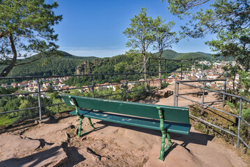  A green wooden bench on a high viewpoint overlooking the town of Dahn, with the town's buildings and the forested hills of the Pfälzerwald visible in the valley below.
