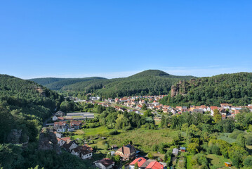 A panoramic view of the town of Dahn in the Pfälzerwald forest, showing the red-roofed buildings nestled in a green valley with a prominent rock formation behind it, under a clear summer sky.