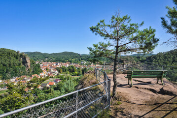  A green wooden bench on a high viewpoint overlooking the town of Dahn, with the town's buildings and the forested hills of the Pfälzerwald visible in the valley below.