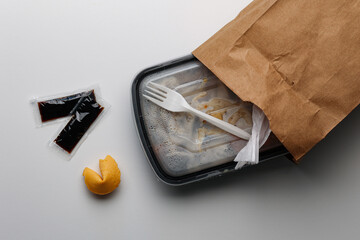 Asian takeout box with plastic fork and paper bag, soy sauce packets, and fortune cookie arranged on white surface.