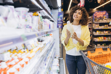 Woman choosing milk in supermarket refrigerator shelf pushing shopping cart