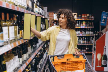 Woman choosing wine bottle from supermarket shelf pushing shopping cart