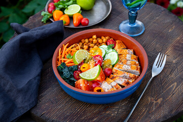 Buddha bowl with baked chicken breast, chickpeas and fresh vegetables on a table in a summer cafe