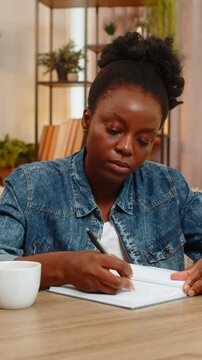 African American young woman sitting at table on home sofa thoughtfully sketches a new logo in notebook, deeply focused on creative design work. Black girl expresses artistic ideas with inspiration.