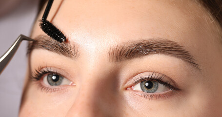 Fototapeta premium Plucking and brushing young woman's eyebrow with spoolie on light grey background, closeup