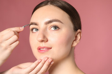 Fototapeta premium Cosmetologist plucking young woman's eyebrow with tweezers on pink background, closeup