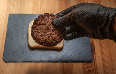 Closeup of hand with black glove placing grilled beef burger patty on slice of bread, perfect for food, cooking, sandwich, and lifestyle concepts.