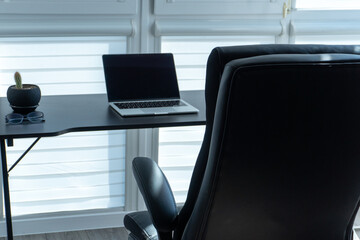 A modern workspace features a sleek black chair, a laptop on a minimal desk, and a small plant, all bathed in natural light from large windows