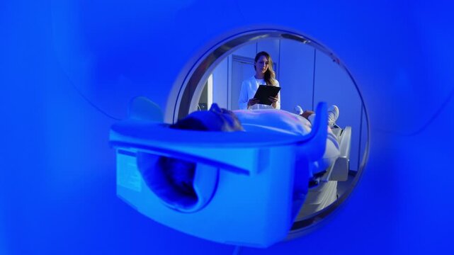 Medical worker standing confidently near MRI tunnel while male patient resting on table. Patient lies on MRI table and undergoes magnetic resonance imaging. Woman in robe and protective cap enters
