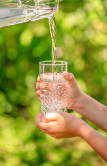 Child hands holding a glass while clean fresh water is being poured outdoors, symbolizing hydration and healthy lifestyle
