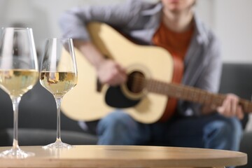 Glasses with white wine on table and man playing guitar indoors, selective focus