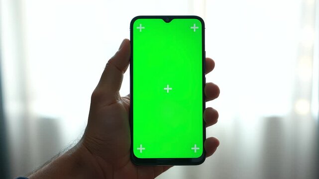 A person holding a modern smartphone vertically, displaying a green screen with tracking markers for easy compositing, perfect for showing an app interface against a bright indoor background
