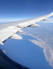 Obraz premium Aerial view from the plane window captures the wing against a clear sky and a coastal view below. Deep blue sea meets a coastline creating a beautiful contrast between water and land from altitude