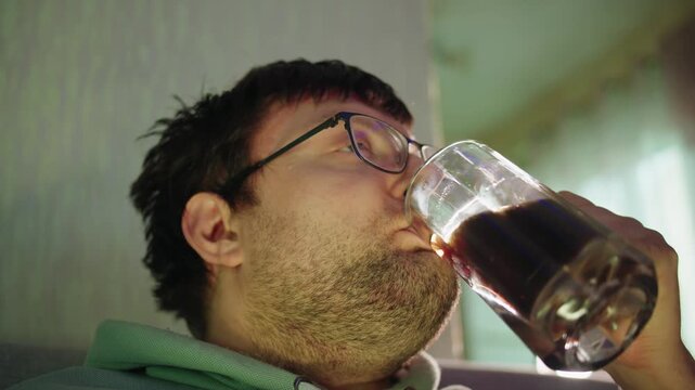 Bottom view of a bearded caucasian adult shaggy man with glasses drinking cola from a glass mug in a living room. Consumption of sugary unhealthy drinks at home. Portrait of the man looking forward