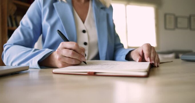 Professional woman writing in notebook seated at workplace desk, working or studying in office, carefully records information, busy in goals or to-do list creation, productivity, workflow organization