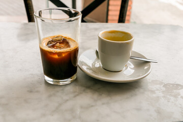 Glass of iced coffee and cup of espresso sit on a marble table at a cafe