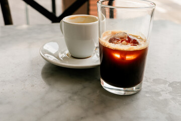 Glass of iced coffee and cup of espresso sit on a marble table at a cafe