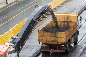 Milling machine working and removing asphalt from the road and pouring it into a truck. Road construction or rehabilitation on a rainy day © Abdull