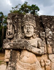 Obraz premium Weathered stone carving of a serene female deity, adorned with elaborate headdress and jewelry, set against a backdrop of lush greenery and cloudy skies