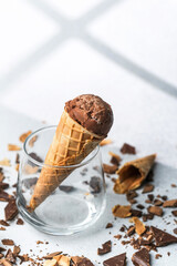 Waffle cone with chocolate ice cream placed in glass surrounded by scattered chocolate chunks on light surface