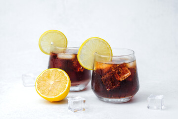 Two glasses of dark soda with lemon slices and ice cubes served on light background with citrus garnish