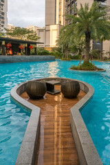 A tranquil poolside patio features two wicker chairs, a small table, and a wooden deck. The pool is surrounded by lush greenery and tall buildings.