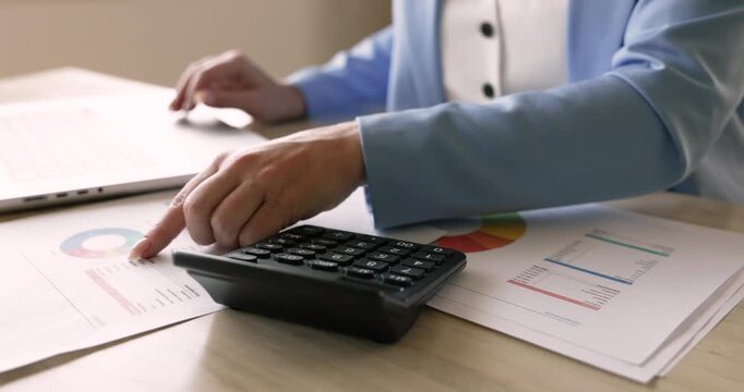 Close up woman using calculator and laptop while reviewing diagrams, charts, calculating company expenses or incomes, preparing budget, working on financial forecast, updating spreadsheet at workplace