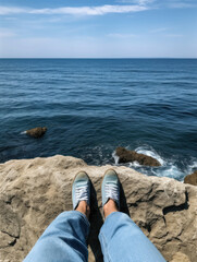 Legs in jeans and sneakers sitting on rocky cliff above calm blue ocean.
