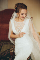 A bride in an elegant white gown with lace details stands in a living room. Warm light and a sunset painting on the wall create a serene and festive atmosphere.