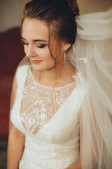 Fototapeta premium A bride in an elegant white gown with lace details stands in a living room. Warm light and a sunset painting on the wall create a serene and festive atmosphere.
