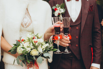 Bride with bouquet and groom in burgundy suit holding wine glasses decorated with red ribbons. Wedding toast detail.
