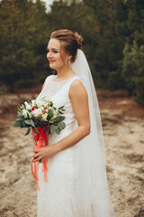 Beautiful bride with a bouquet posing by the water, her veil flowing in the wind, creating a tender and romantic scene.