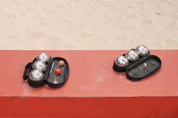 Two petanque sets in black cases with metallic boules and red target balls placed on red concrete edge near sand terrain, evoking relaxed outdoor sport in city environment