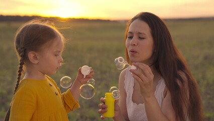 Fototapeta premium Happy Mother and her little Daughter playing together outdoor, blowing soap bubbles, having fun on backyard. Nature. Beauty Mum and her Child in Park together. Family, Mother's Day Joy. Mom and Baby