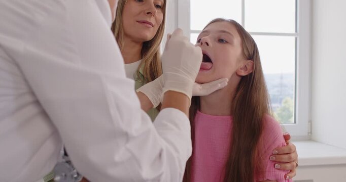 Female doctor in gloves performs a throat swab exam for a child, while the mother stands close in a bright clinic. Doctor swabs child's throat during exam. Caring pediatric visit builds trust.