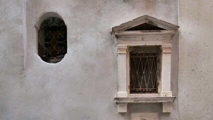 Weathered Venetian wall featuring two contrasting windows—one oval with a wrought iron grille, the other rectangular with a triangular pediment and diamond-patterned bars. A study in texture, age, and