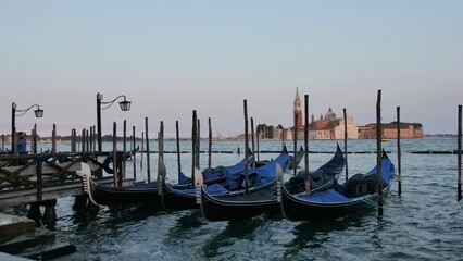 Gondolas docked in Venice with a gondolier steering across calm waters. In the background, the Church of San Giorgio Maggiore rises with its bell tower&mdash;capturing the serene and iconic spirit of the ci