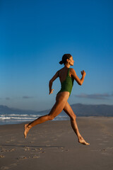 Fit young woman in green swimsuit mid-air as she jumps gracefully along the beach at sunrise, showing strength, energy, and freedom under a clear blue sky.