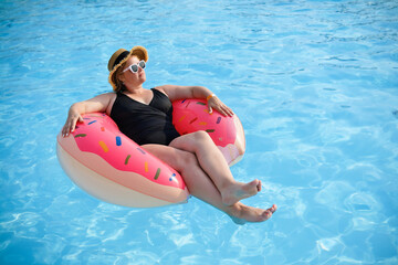 Beautiful white European woman in black swimsuit wear hat and sunglasses relaxing in swimming pool with rubber ring.