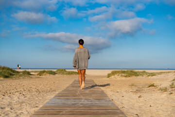 Woman in oversized sweater walking barefoot on a wooden path toward the beach, peaceful morning scene with cloudy blue sky and distant ocean horizon.