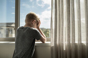 Sad unhappy lonely little boy at home crying next too window 